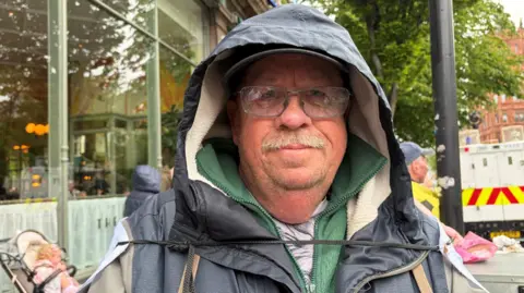 A man with clear spectacles and a grey moustache stairs to the camera. He wears a navy waterproof jacket, with a green fleece underneath. Over his left shoulder is a white police van. Over his right, a baby sits in a cream pram below a clear window. 