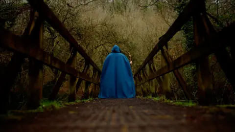 Cherry Tree A person, standing on a bridge facing away from the camera, wearing blue robes and holding a wooden staff. Trees can be seen in the background.