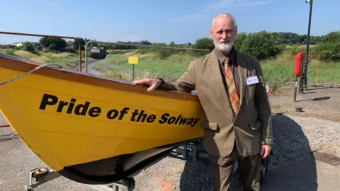 A man with white hair and a beard, wearing a brown suit and an orange tie, stands next to a yellow rowboat with "pride of the Solway" in black lettering on the side.