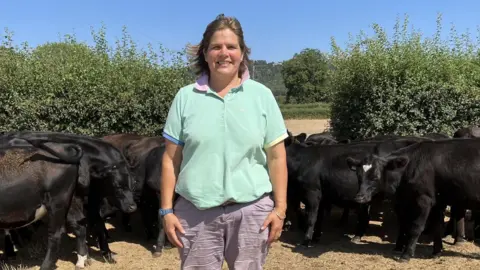 Leanne Rinne/BBC A woman in a mint green polo shirt standing in a field in front of a heard of black cows