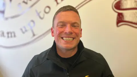 A man with short brown hair looking at the camera. He is giving a toothy smile and wearing a black zip jacket.