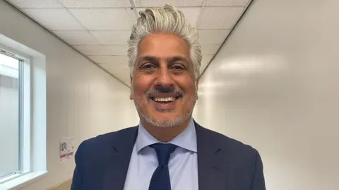 Chohan pictured looking into the camera and smiling. He has short silver hair, brushed into a quiff, and a short beard. He's wearing a navy blue blazer, navy tie, and a light blue collared shirt. He's stood in a long hospital corridor, which has white walls and a white ceiling. 