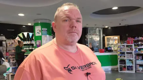 Christopher Connor stands in a pharmacy. He is wearing an orange t-shirt with black writing that says 'silverback'. He has grey hair and blue eyes.