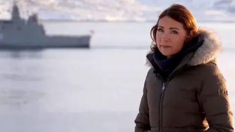 Katya Adler in a coat by the sea with a boat in the background.