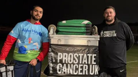 Two men next to a silver case full of football equipment. There is a banner reading Prostate Cancer UK. the man on the left has a sky blue shirt with Alder Hey written on. He has a red undershirt. The man on the right is wearing a black hoodie with Prostate Cancer UK on it in white lettering