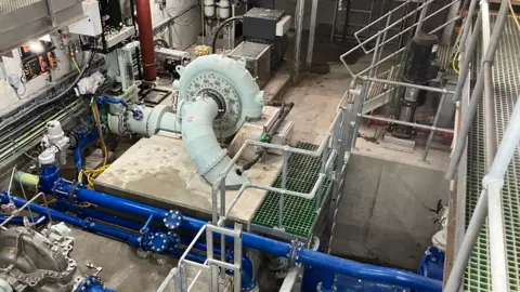 A view inside an industrial building, which is full of brightly coloured pipes, a turbine and other kit associated by hydro power generation 