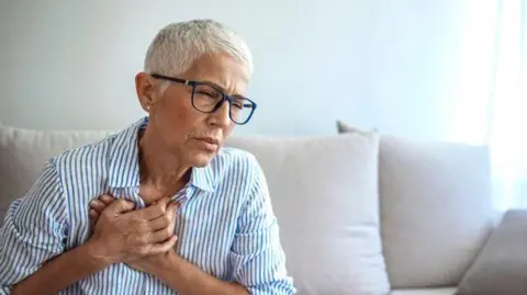 A woman with short grey hair is sitting on the sofa and is pressing both her hands to her chest. Her facial expression suggests she has pain in her chest. She has glasses on and is wearing white shirt with blue stripes. 