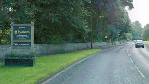 A Google Streetview screenshot of the road into Hexham, with a green sign welcoming people to the town, mounted on a wooden frame with a planter at its base.