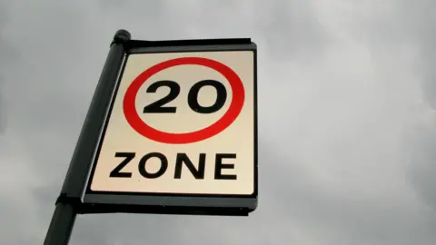 A road sign with the number 20 and the word "zone" in black writing on a white background. There is a red circle around the number, and a cloudy sky in the background.