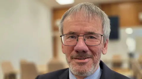 Paul Moseley/BBC Brian Watkins is wearing glasses. He has a navy suit jacket over a blue and white striped shirt with tie.