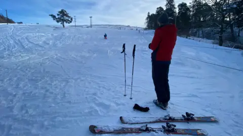 A man stands looking at a slope of snow - with skis at his feet.