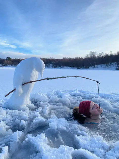 Fiona Clarke Woman submerged in a frozen loch, holding a fishing rod in her mouth which is held by a 'snowman' penguin.