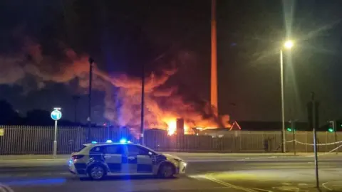 Johnny Greenwood Scene of the fire. A police car with blue lights flashing, with smoke and flames seen in the background.