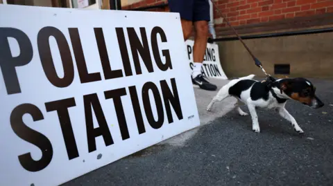 Small black and white dog strains on the lead outside a polling station, which has a large sign outside