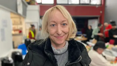 Michelle Swift, a woman with blonde hair, is looking at the camera and smiling. She is wearing a black and white striped jumper and a black coat over the top. Behind her is a group of people taking part in a cookery session.