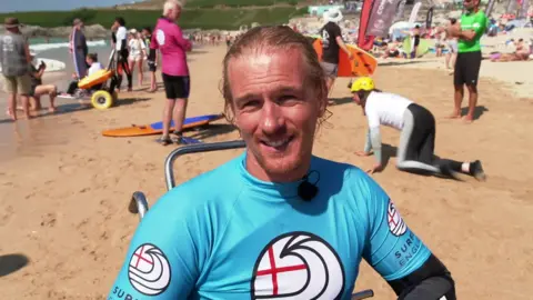 Cam Treble wears a blue Surfing England top over a wetsuit and smiles into the camera 