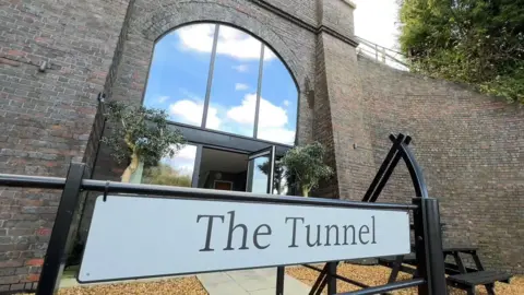 A former railway tunnel. The arch is filled with large panes of clean glass and there is a patio in front of it. At the front is a sign that says 'the tunnel'.