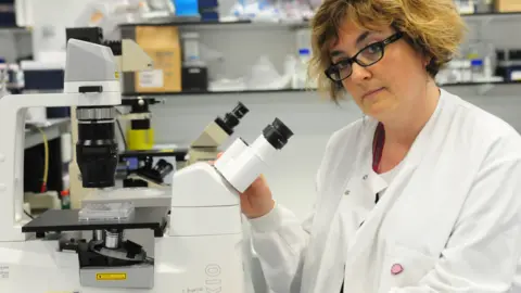 Brain Tumour Research A woman in a lab coat and glasses stood next to a microscope. 