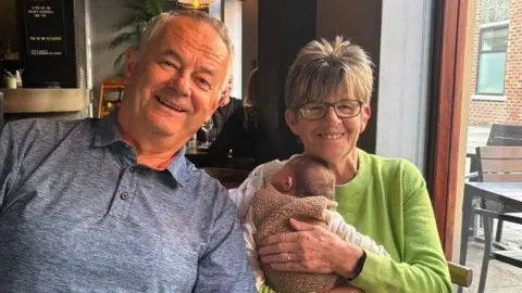Left to right: Tad with grey hair wearing a grey polo top sitting alongside Nicki Detko with short grey and blond hair holding one of her grandchildren who is sleeping wrapped in a blanket. Nicki is wearing a lime green jumper. They are sitting in a restaurant smiling.