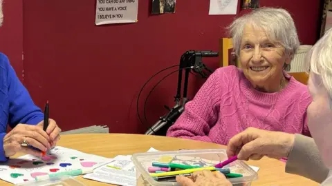 BBC Pensioner Mary is at a table where others are colouring in with felt-tip pens. She has grey hair and is wearing a pink jumper. She is smiling at the camera and is sitting with others who are also colouring in with felt tip pens. 
