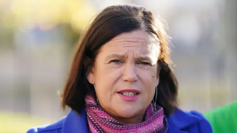 Pa Media Mary Lou McDonald - Woman with brown hair with a shoulder length staring while speaking. She wears a pink scarf and a blue coat.