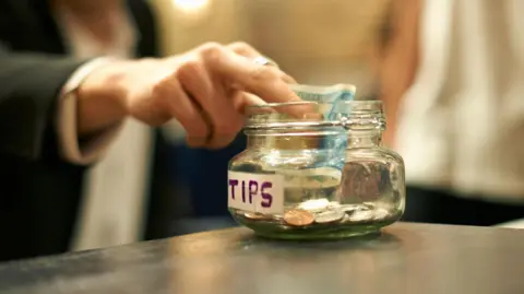 Getty Images A man in a black and white suit places a five pound note into a clear tip jar with 'Tips' written on it. The jar sits on a black counter