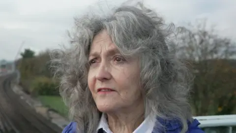 BBC/Nathan Turvey Darlene, a woman with long, curly grey hair wearing a bright blue puffer jacket over a white collared shirt.