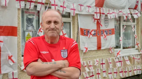 Harriet Morter / BBC Paul Bibby wearing a football top standing in front of his home, which is covered in England Flags