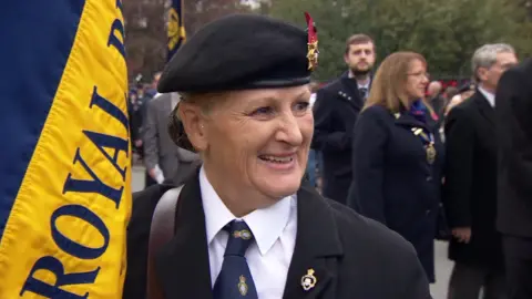 Kate holds her yellow standard and is dressed in black.  She is smiling at the interviewer.