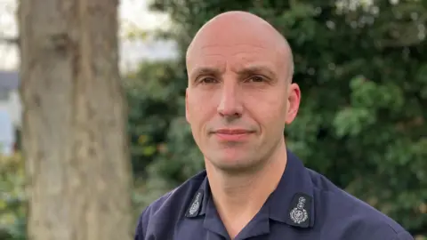 Rick Hylton wearing a navy shirt in front of a tree and a bush. He has a shaved head and looks serious.