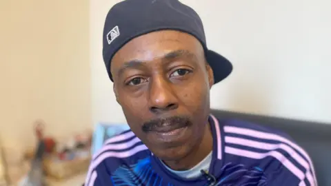 A man wearing a black baseball cap turned backwards wearing a blue jumper with white stripes along the shoulder and a white t-shirt underneath it