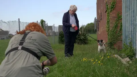 Kay Sebern outside a polling station with her Jack Russel Sasha - Kay has a navy blue top and trousers on with a light blue shirt underneath. Sasha is black and white and being held on a retractable leash. 