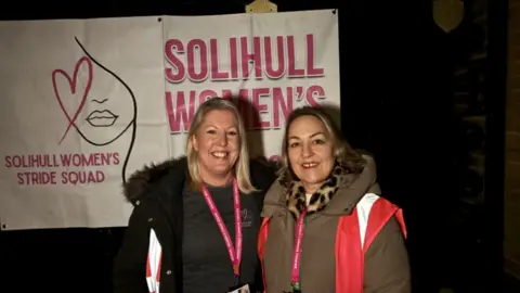 Two women smiling into a camera. They have both got pink high-vis jackets on and coats. They have got pink lanyards on and are smiling. One has blonde hair and one has brown hair. 