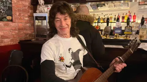 A man with a long mullet holding a guitar in a pub. He has a white top on and is vaguely smiling at the camera. He's stood besides the bar. 