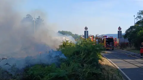 Kieran Moyes Two fire engines, parked in the middle of a road curving through the countryside, spraying water across a patch of burning brush. Orange flames glimmer through the plume of steam.