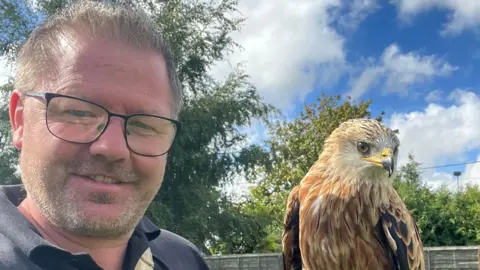 Mark Birdsall taking a selfie outside with his red kite, Rusty. He has short, brown hair and short, greying stubble. He is wearing a blue polo shirt and black-rimmed glasses. Rusty has golden feathers and a yellow beak.