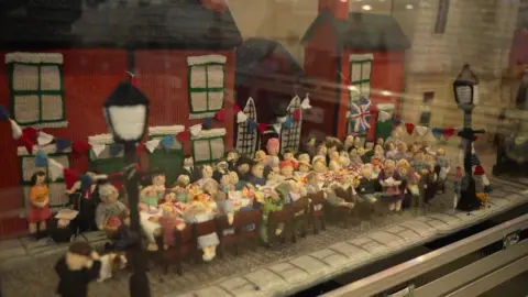 A yarn depiction of a large street party. Dozens of people are gathered around tables outside, with bunting on houses in the background.