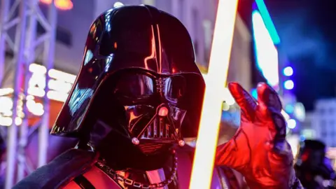 An actor dressed as Darth Vader holds a glowing lightsabre outside a London theatre