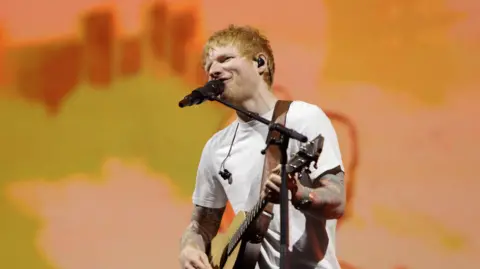 A red-haired Ed Sheeran sings into a microphone while playing a guitar. He is wearing a white tee-shirt