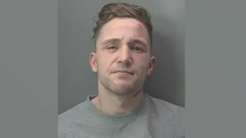 Cambridgeshire Police A police mug shot of Dillon Currie aka Abeeb Hussain. He has light brown hair which is longer on the top and very short on the sides and has subble. He is wearing a grey sweatshirt.  