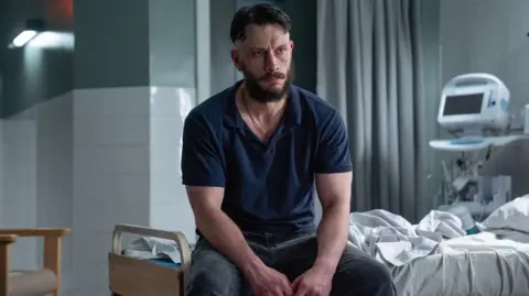 BBC Actor Richard Gadd in a hospital scene in his new series Half Man - he sits looking morose on a dishevelled hospital bed, wearing a blue polo shirt and jeans. He is muscular and has dair hair, a beard and a moustache.