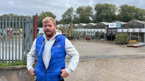 Man who runs the Maulden Garden Centre is standing by the entrance to the site with the flooded area behind him. He is wearing a white hooded jumper under a blue sleeveless jacket.