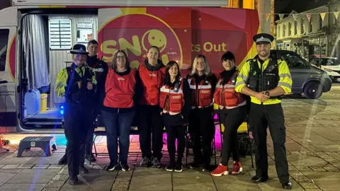 Five women in red jackets stand in front of a white and red van. Three police in high visibility jackets stand next to them in a town centre. 