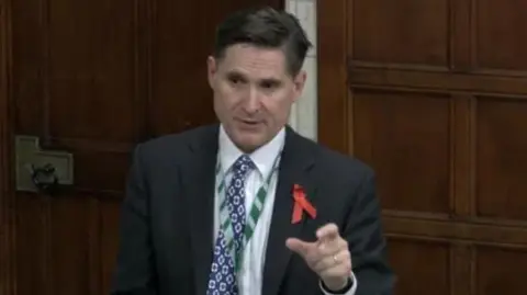 UK PARLIAMENT The MP Charlie Maynard speaks in Parliament. He is wearing a darn suit with a white shirt and blue patterned tie. The walls behind him are large oak wood panels and he is raising his hand to make a point