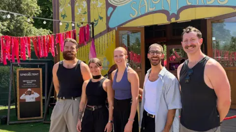 James McConnell/BBC Sam, on the left, stands head and shoulders above Christina who is next in line. Next in line stand Chelsea, Jon and Nathan. All five differ in height and build, but are dressed similarly for the hot weather in vests and leotards. They are smiling at the camera, and surrounded by festival decorations of bright pink bunting and festoon lights. 