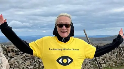 Contributed Dawn Blezard smiles at the camera at the top of a mountain. She has blonde hair and wears sunglasses as well as a black long sleeved top with a yellow T-shirt over the top. She has a hood over her head.