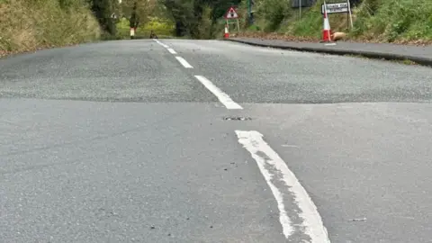 The tarmac and white lines of a roadway, some traffic cones in the distance with a kerb to the right.