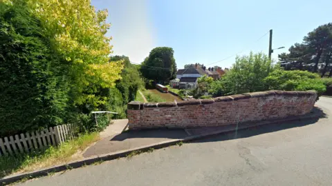 A street view image of a brick bridge over a canal with a barge moored on the water next to a house in the background.