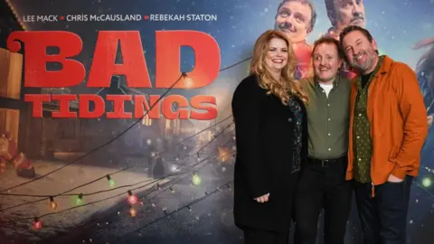 Getty Images A woman in a black coat, a man in a green shirt and a man with a orange coat stand in front of a Christmas themed promotional banner. 