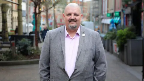 reThINK PR & Marketing Andrew Douglas pictured on a street in Swansea wearing a grey blazer and lilac purple shirt. He is bold and has a dark grey beard.